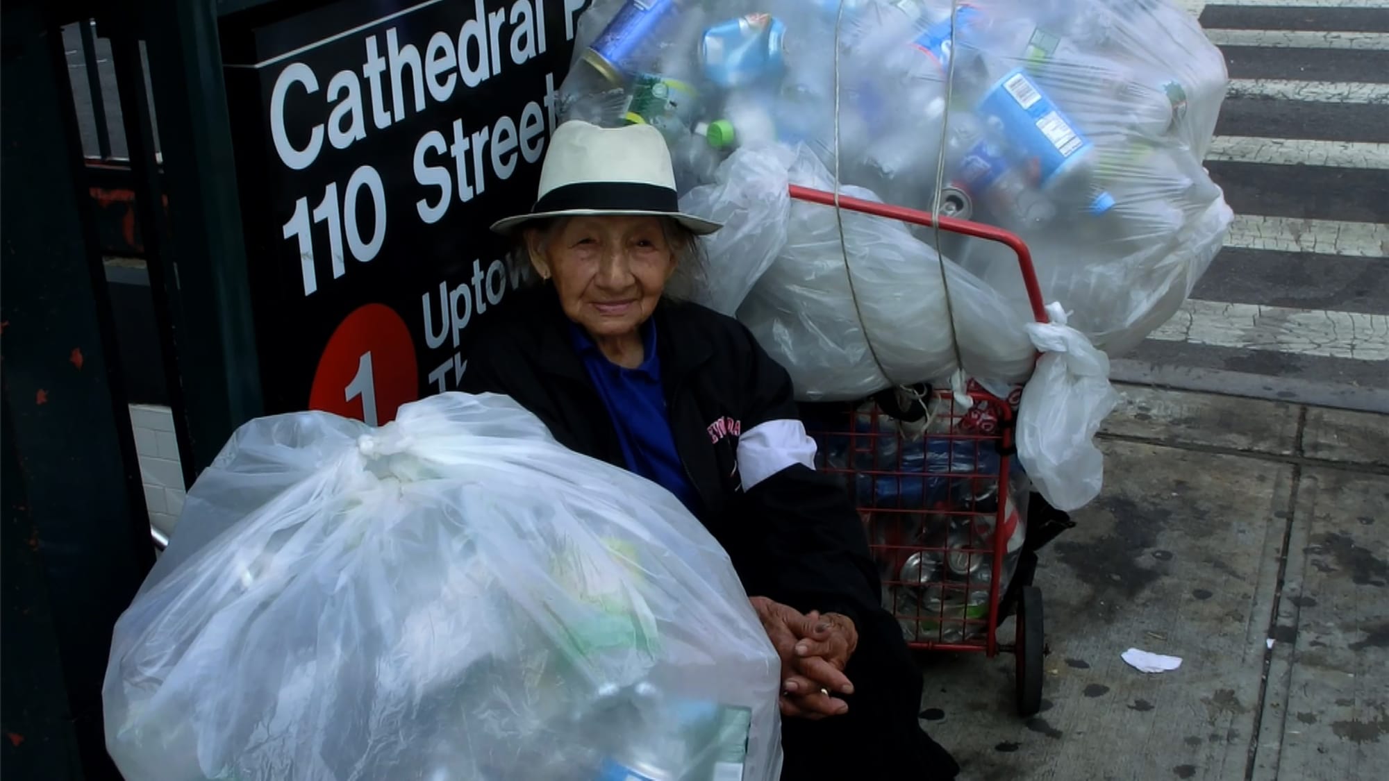 Elderly Asian woman wearing a brimmed hat sits on the street against a subway station sign flanked by two large garbage bags full of cans