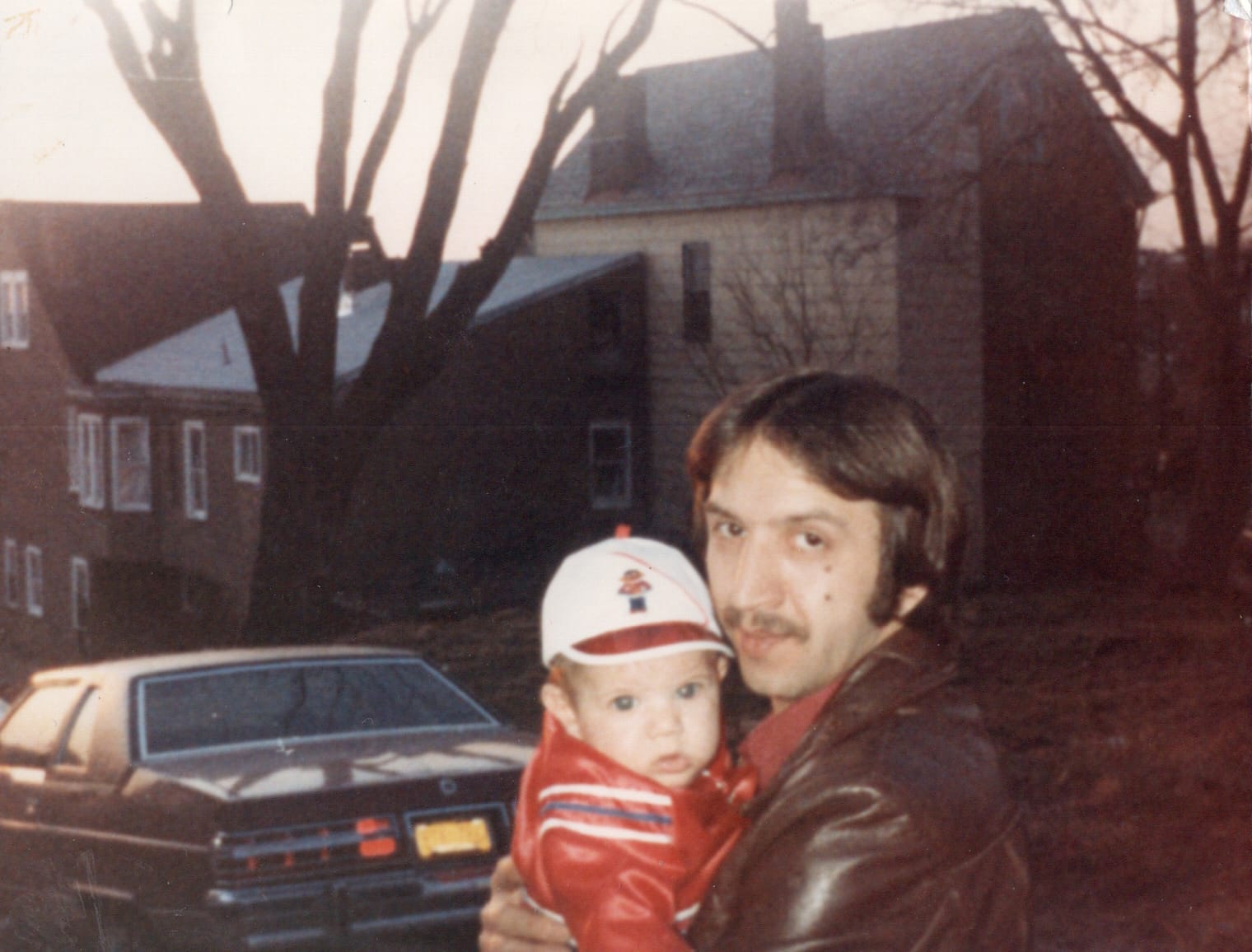 man in brown leather jacket holding a baby in a red leather jacket and white hat