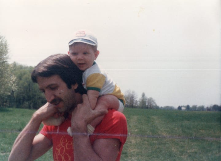 man in red t-shirt carrying a baby on his shoulders through a field