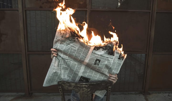 person sitting backwards in a wooden chair reading a newspaper that's on fire